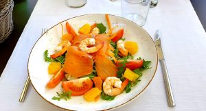 A shrimp and salmon salad with tomatoes and greens on a white plate on top of a white tablecloth.