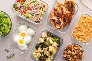 Meal prep with glass containers holding individual meal items including hard boiled eggs, chicken breasts, tabouli, rice, an assortment of nuts, chopped lettuce and broccoli with cauliflower.