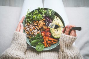 A lady wearing white pants and a cream colored sweater seated on a white couch. You only see her hands holding a fork over a large bowl of veggies that she is holding on her lap.
